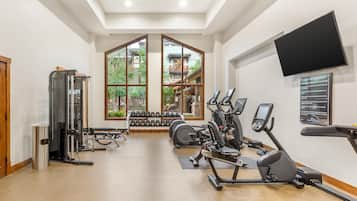 Een fitnessruimte met trainingsapparatuur, een aan de muur bevestigde televisie en grote ramen met uitzicht op een gebouw.
