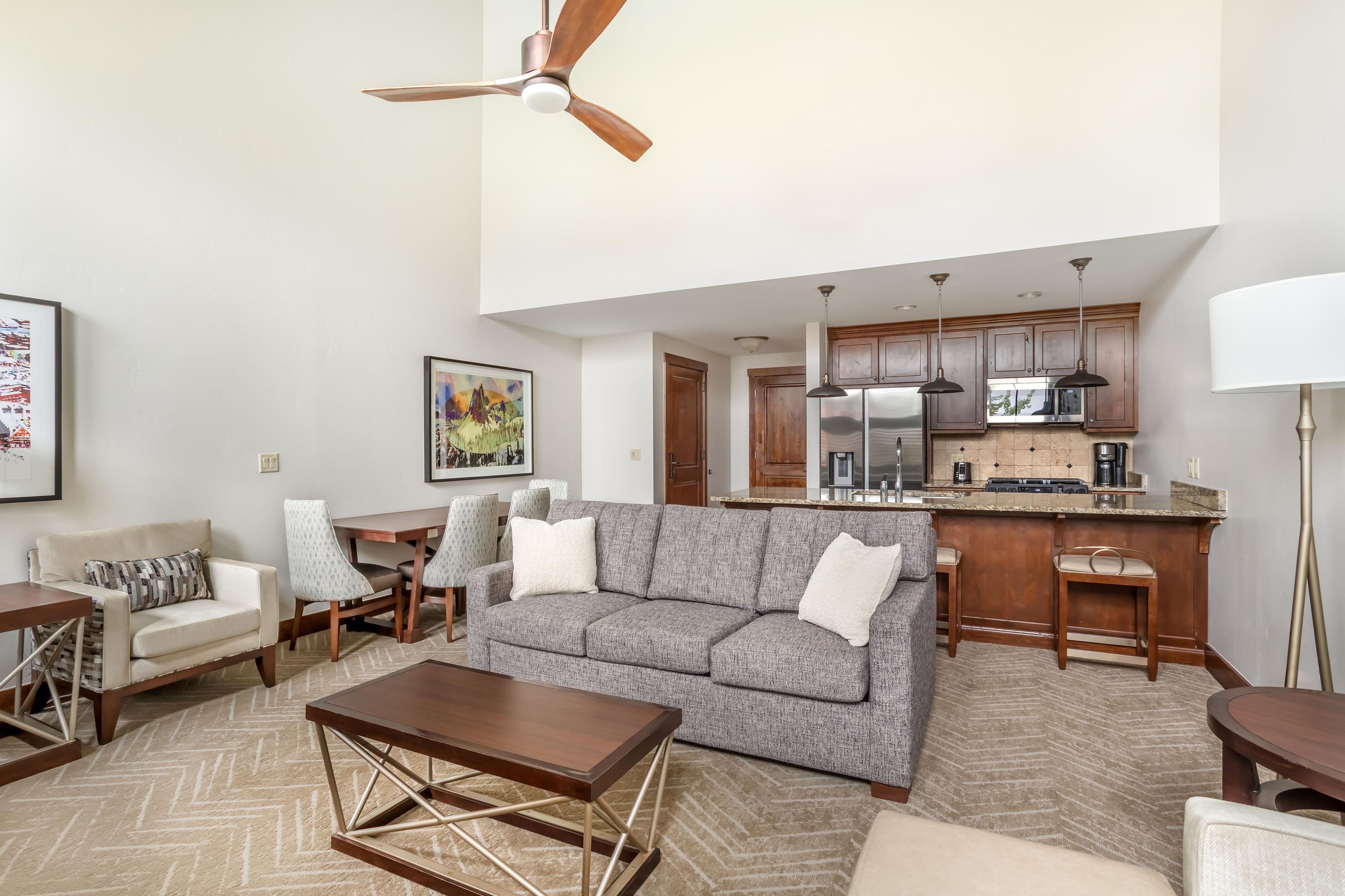 A modern living room with a sofa, coffee table, and chairs. A kitchen with wooden cabinets and appliances is visible in the background.
