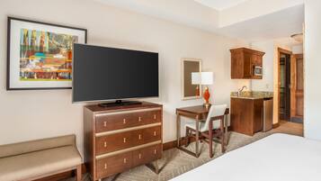 A hotel room with a bed, a desk, a television, a chair, a dresser, a painting, and a kitchen area.