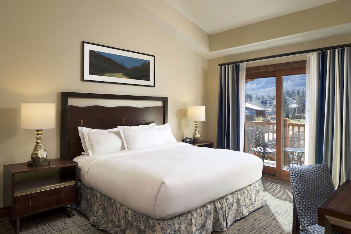 A hotel room with a large bed, two bedside tables with lamps, a framed picture on the wall, and a balcony with a view.