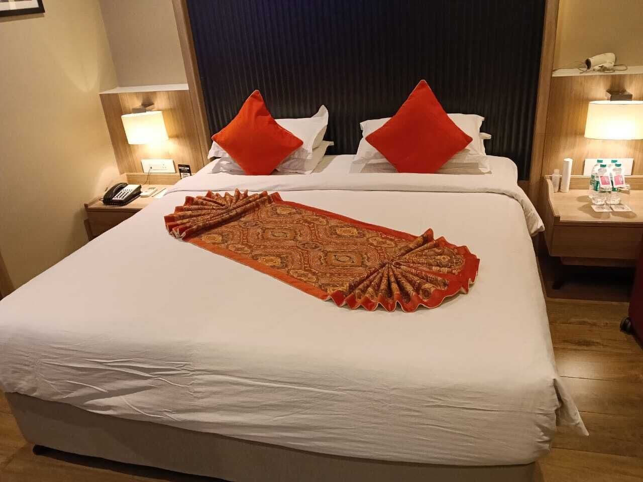 Habitación de hotel con una cama grande, dos almohadas rojas, un plaid estampado, lámparas de noche, un teléfono y una botella de agua en la mesita de noche.