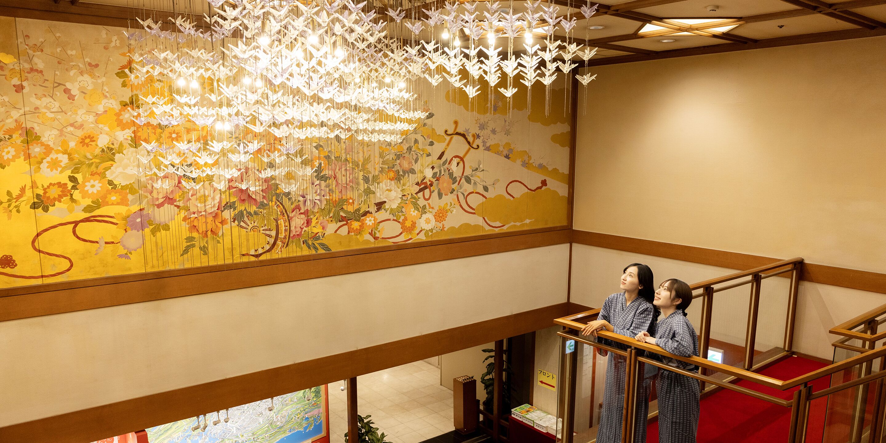 L’intérieur d’un hôtel, avec un grand mur décoré d’une fresque florale, un escalier en bois et un lustre moderne.