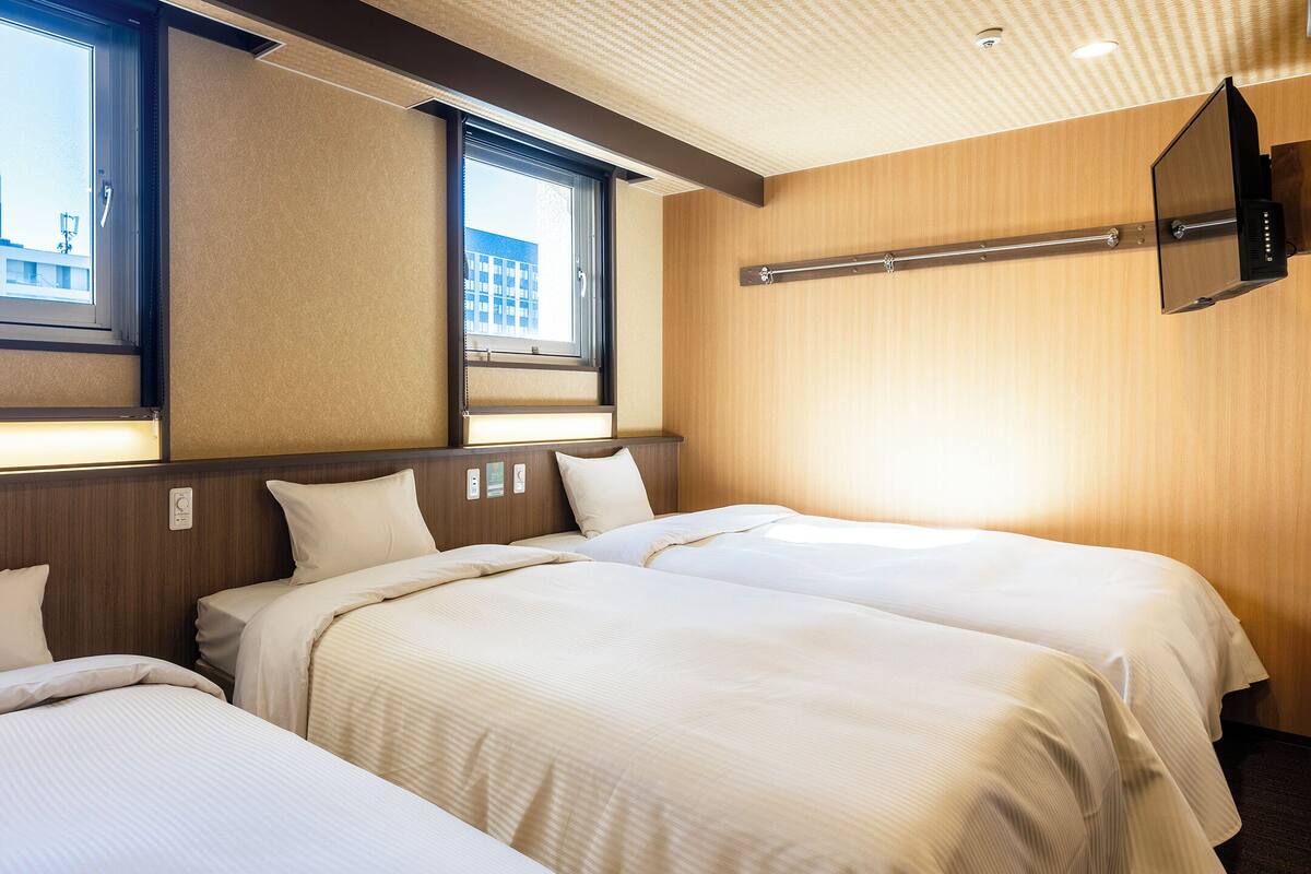 Habitación de hotel con dos camas, un televisor montado en la pared y ventanas con vistas a la ciudad.