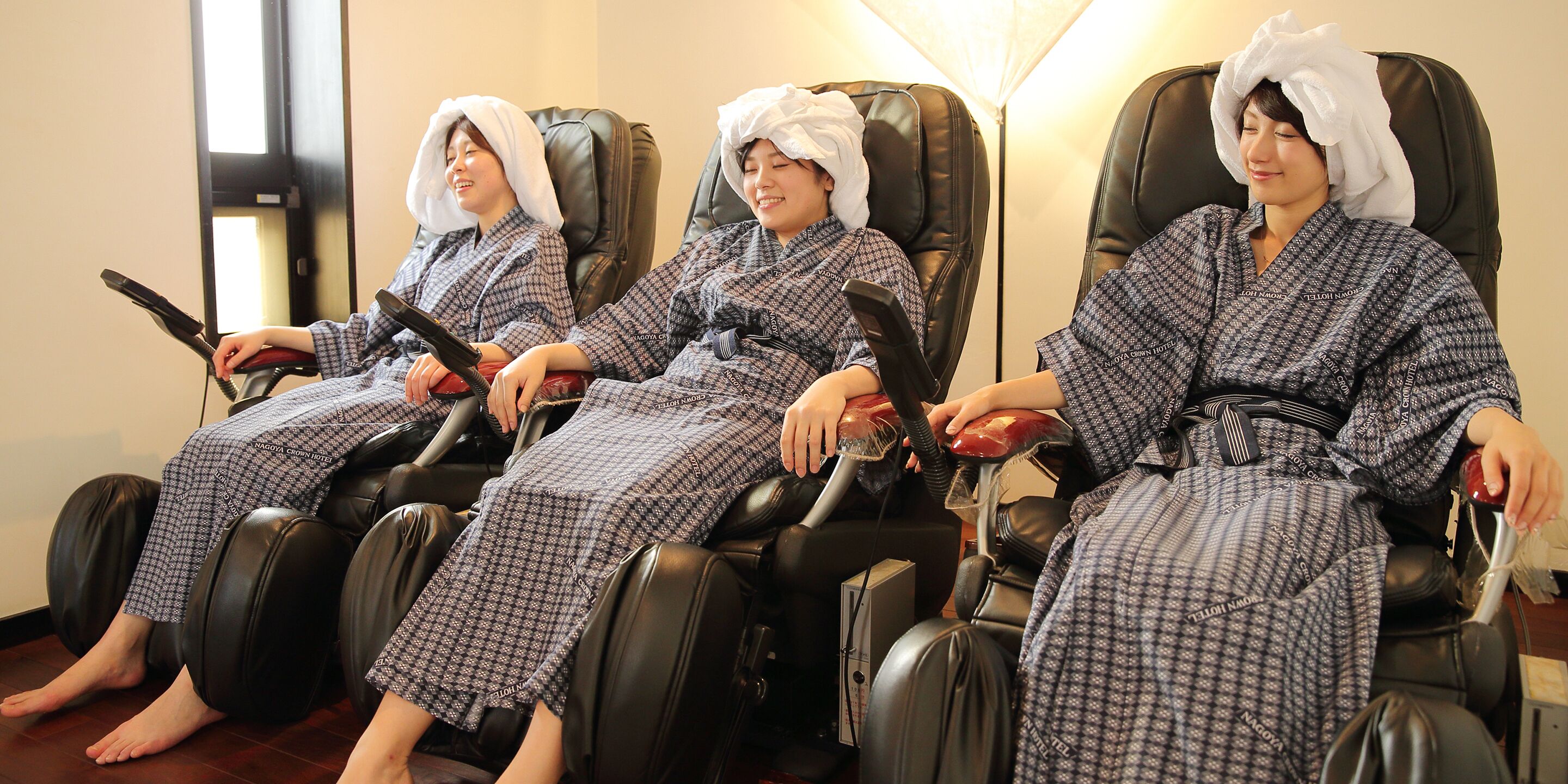 Three individuals seated in a room with massage chairs, each wearing patterned robes and towels on their heads.