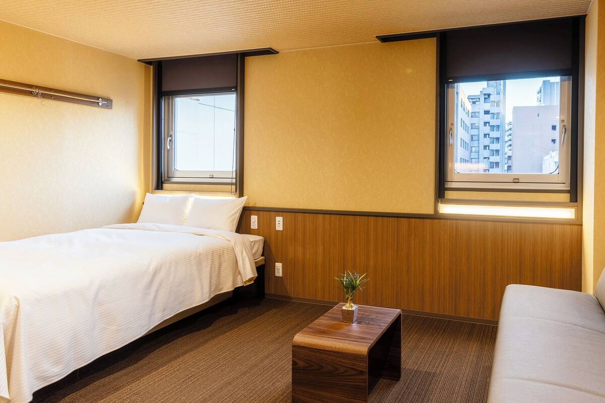 Una habitación de hotel con dos camas, una mesita de madera y vistas a la ciudad a través de dos ventanas.