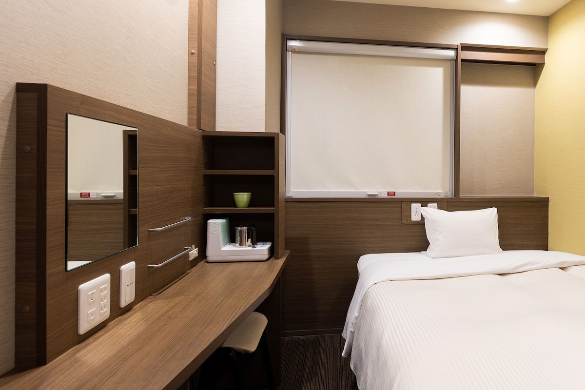 Habitación de hotel compacta con una cama, una mesita de noche, un espejo, un estante con artículos de aseo y una ventana con persianas.