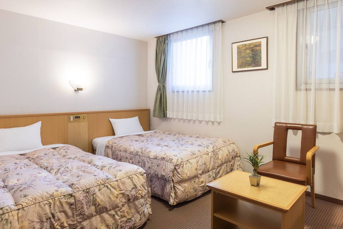 Habitación de hotel con dos camas, una mesita con una planta, una silla y un cuadro en la pared.
