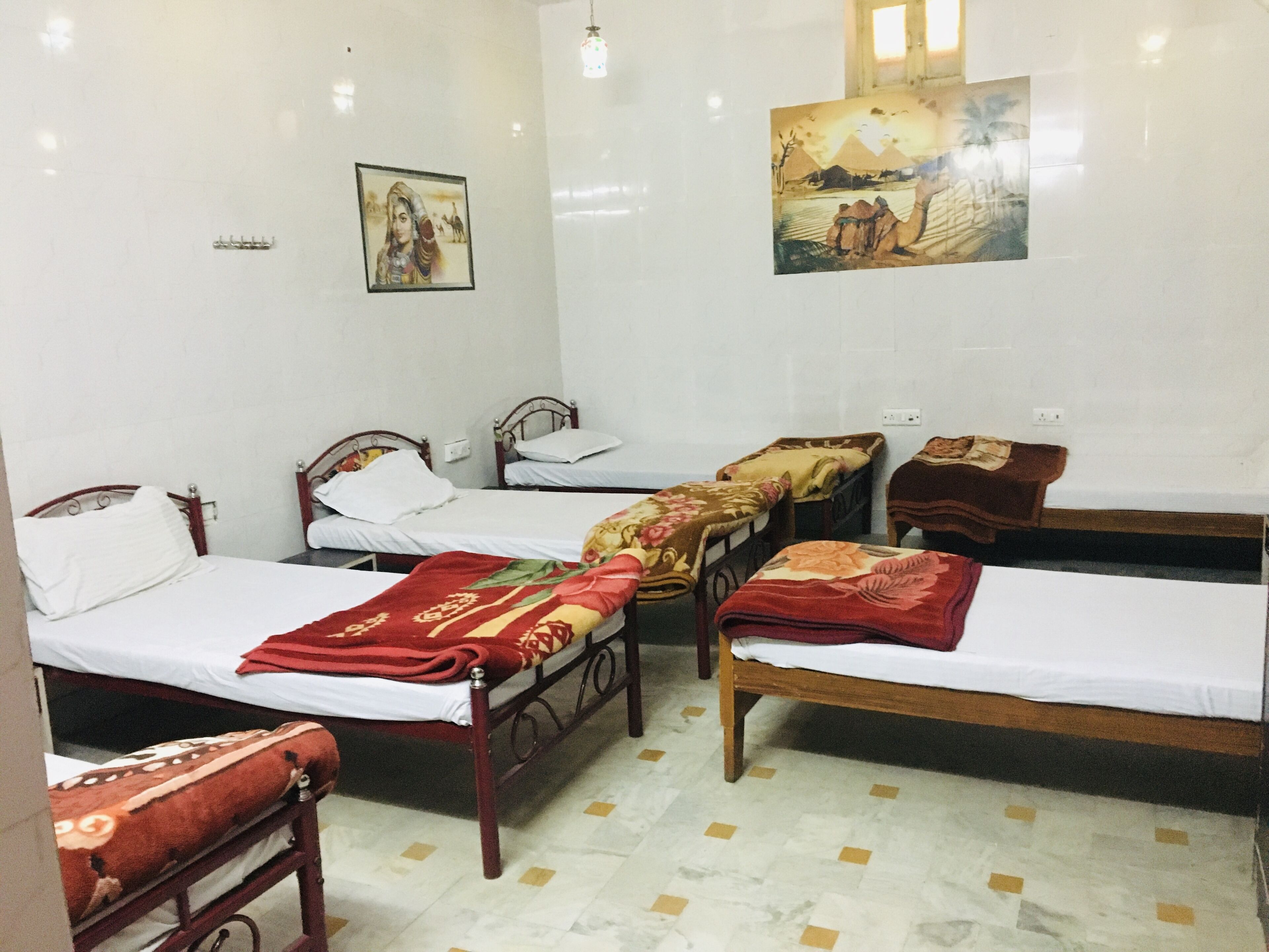 Une chambre avec quatre lits simples, chacun recouvert d’une couverture à motifs différents. On y trouve des appliques murales et deux tableaux encadrés.