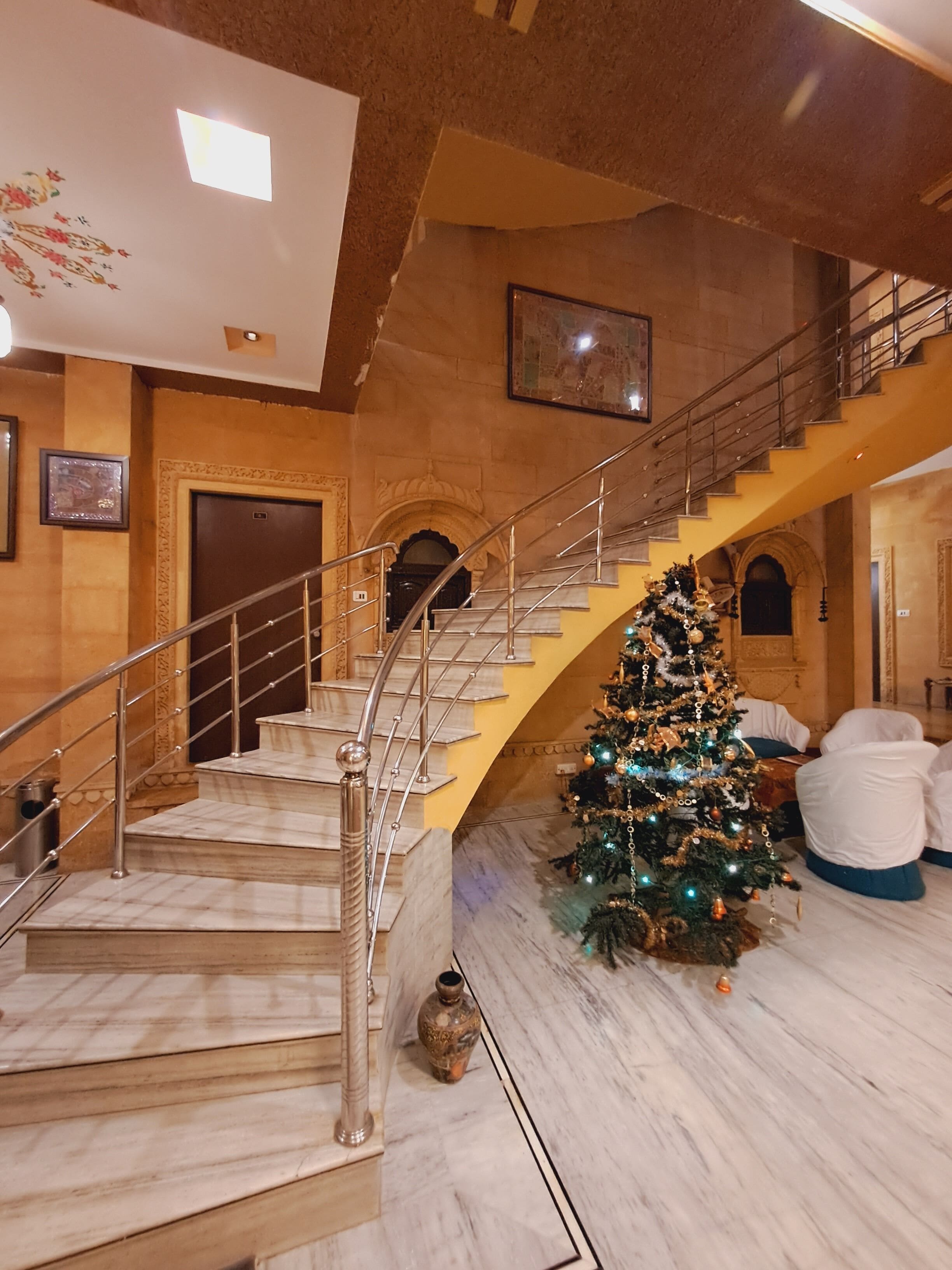 A grand staircase with marble steps and metal railings, leading to a second floor. A decorated Christmas tree is placed on the lower floor near the staircase.