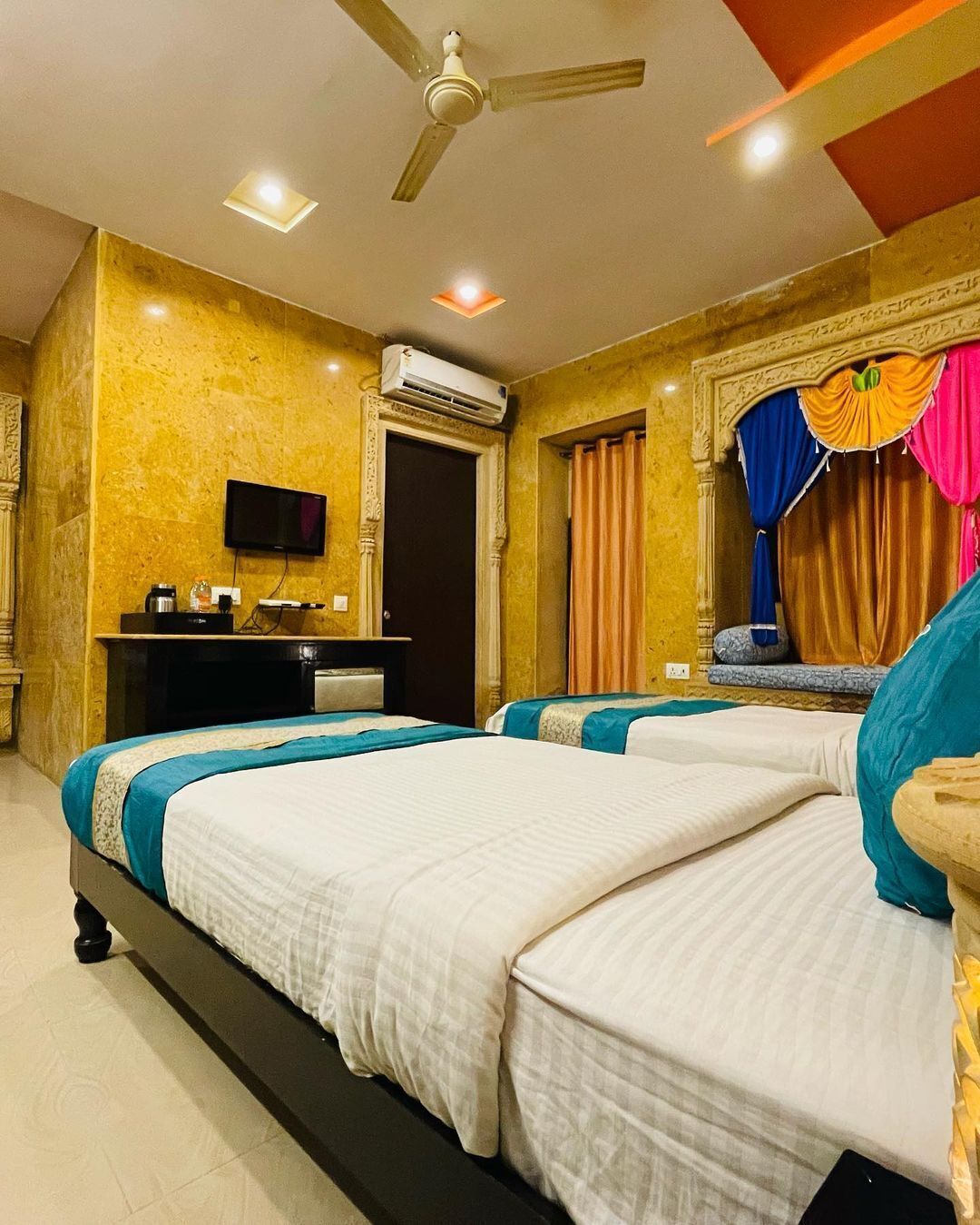 A hotel room with two beds, a desk with a flat-screen TV, an air conditioning unit, and a ceiling fan.