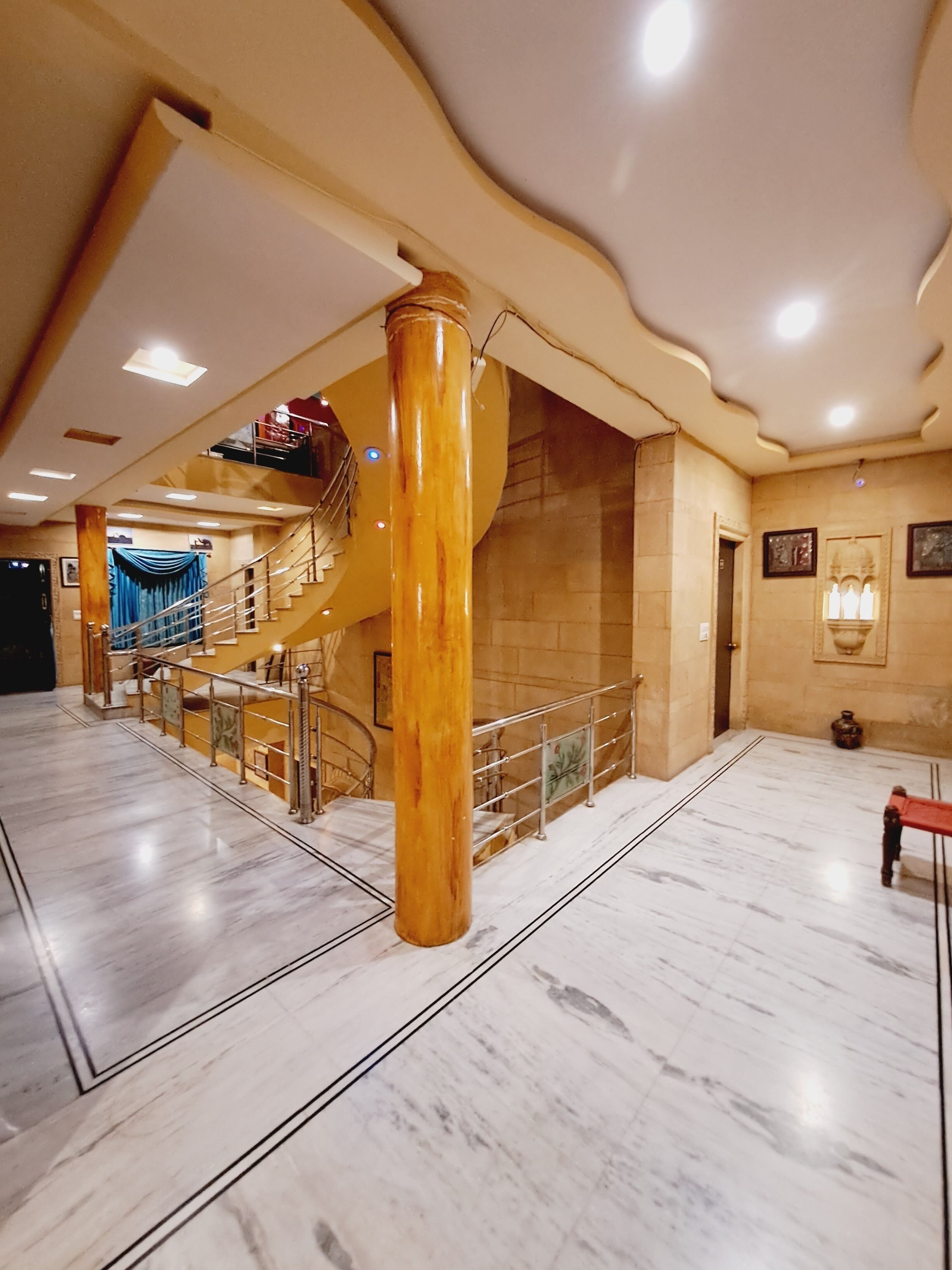 A spacious lobby with marble flooring, a central wooden column, and a staircase with a glass railing.