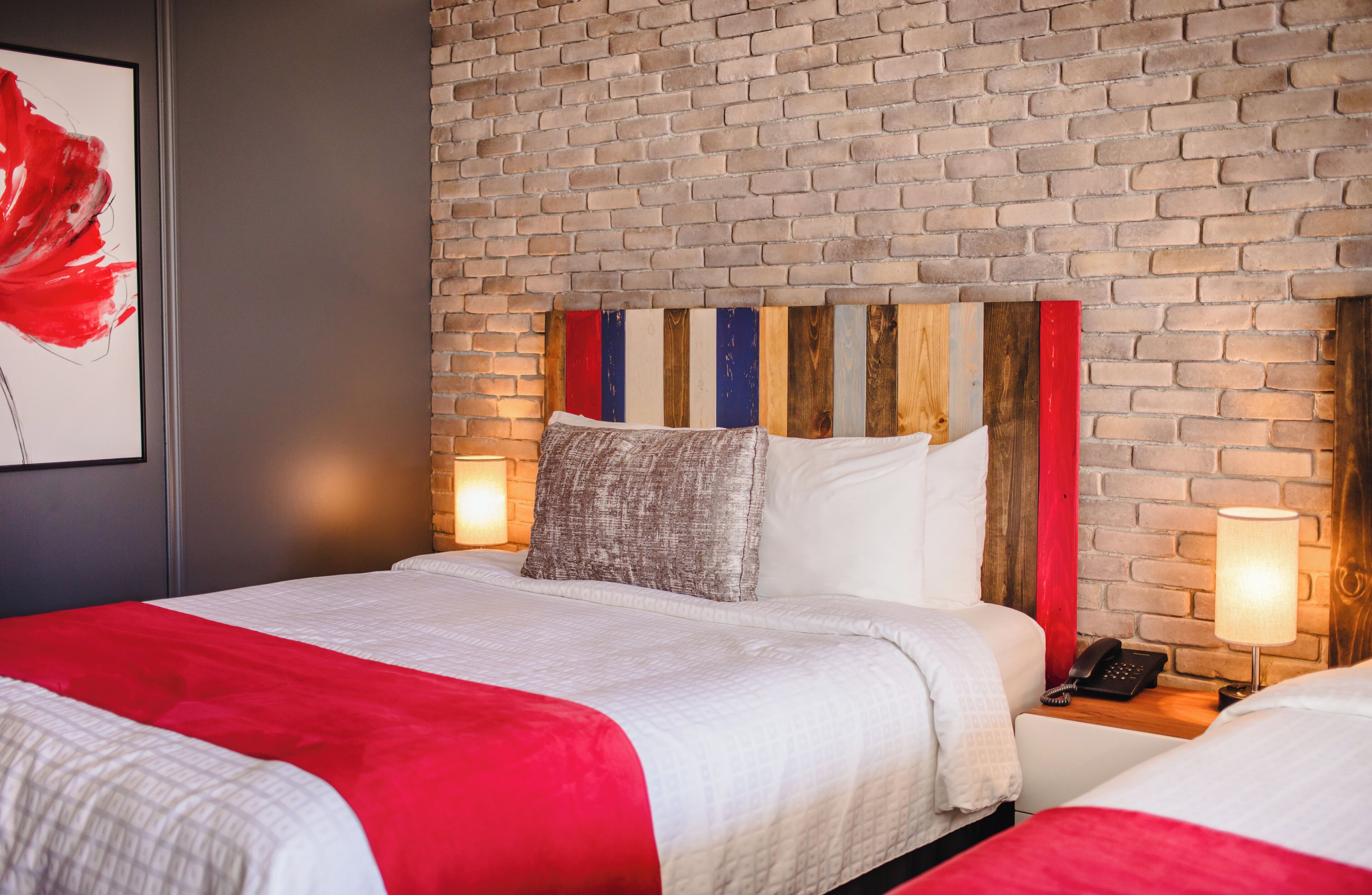 Une chambre d’hôtel avec un mur en briques, un lit avec une literie blanche et un élément décoratif rouge, une table de chevet avec une lampe, et une œuvre d’art encadrée accrochée au mur.