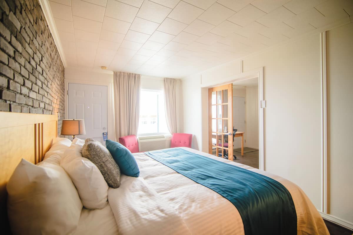 Habitación de hotel con una cama grande, una pared de ladrillo, una ventana con cortinas y una puerta que da a otra habitación.
