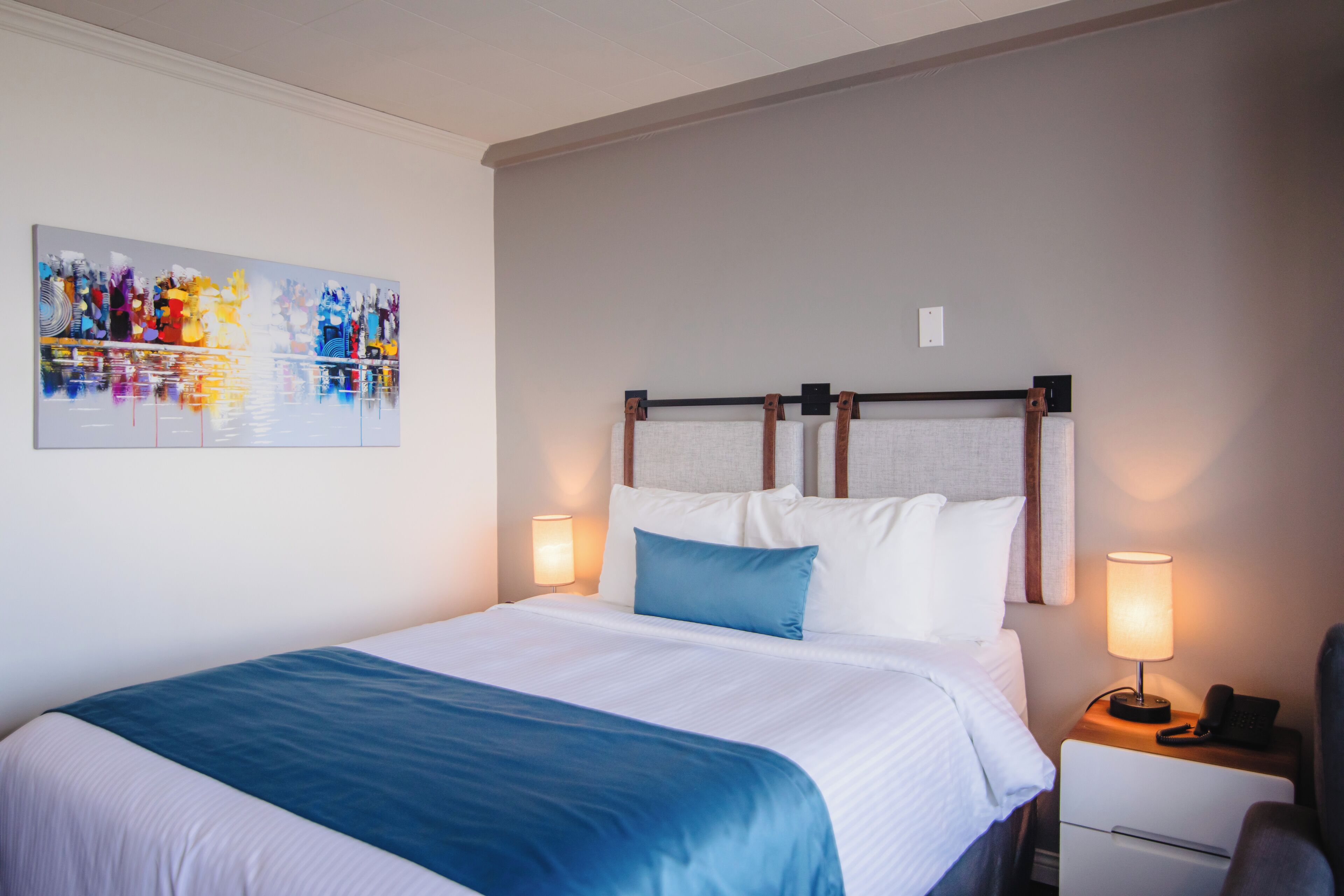 A hotel room with a bed, two bedside lamps, a nightstand, and a painting on the wall.