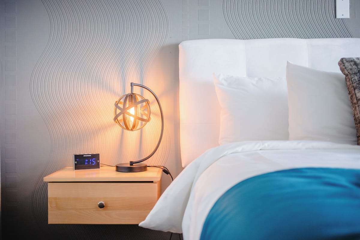 Una cama bien hecha con sábanas blancas, una mesita de noche de madera con un reloj digital y una lámpara geométrica original.