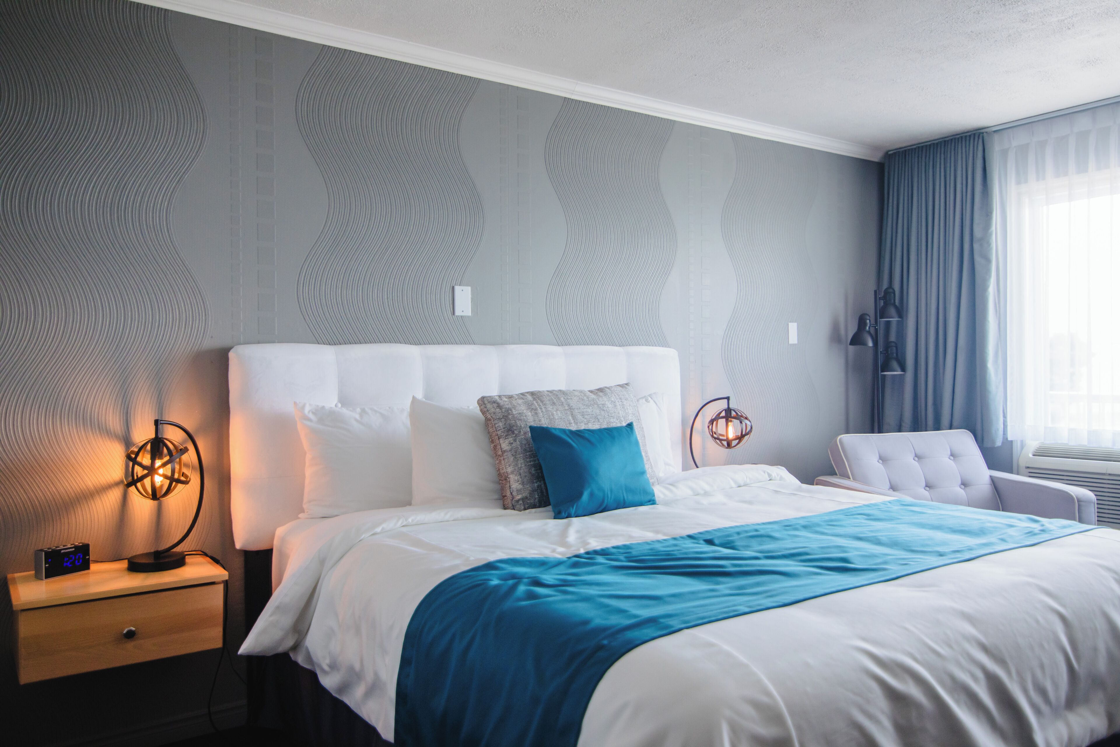 A neatly made bed with a blue throw blanket, a bedside lamp, and a digital clock on a wooden nightstand.