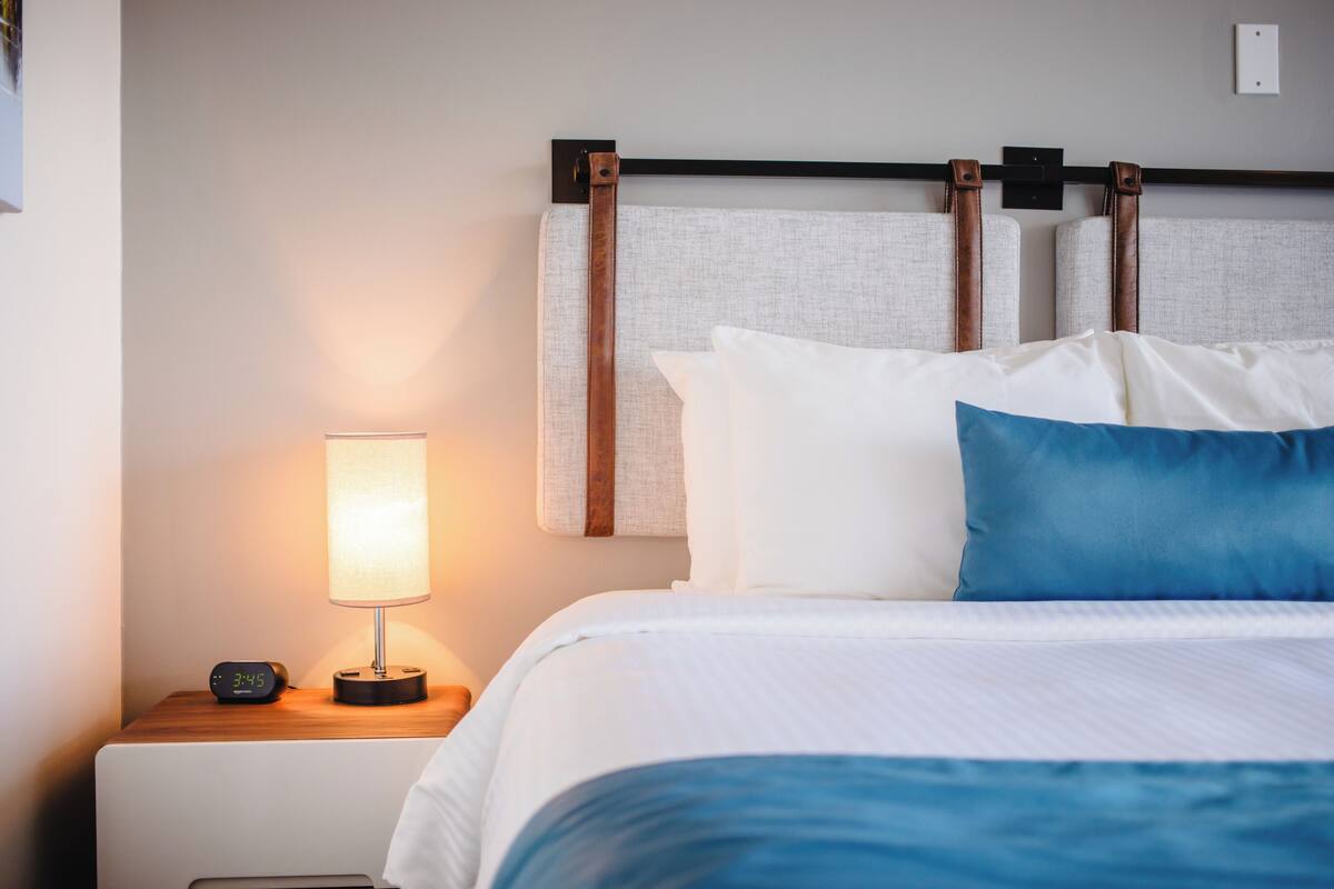 Una cama bien hecha con ropa de cama blanca y azul, una lámpara de noche y un reloj digital sobre una mesita de noche de madera.