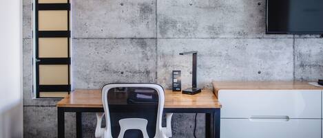 A modern room with a concrete wall, a wooden desk, a white chair, a black lamp, and a TV mounted on the wall.