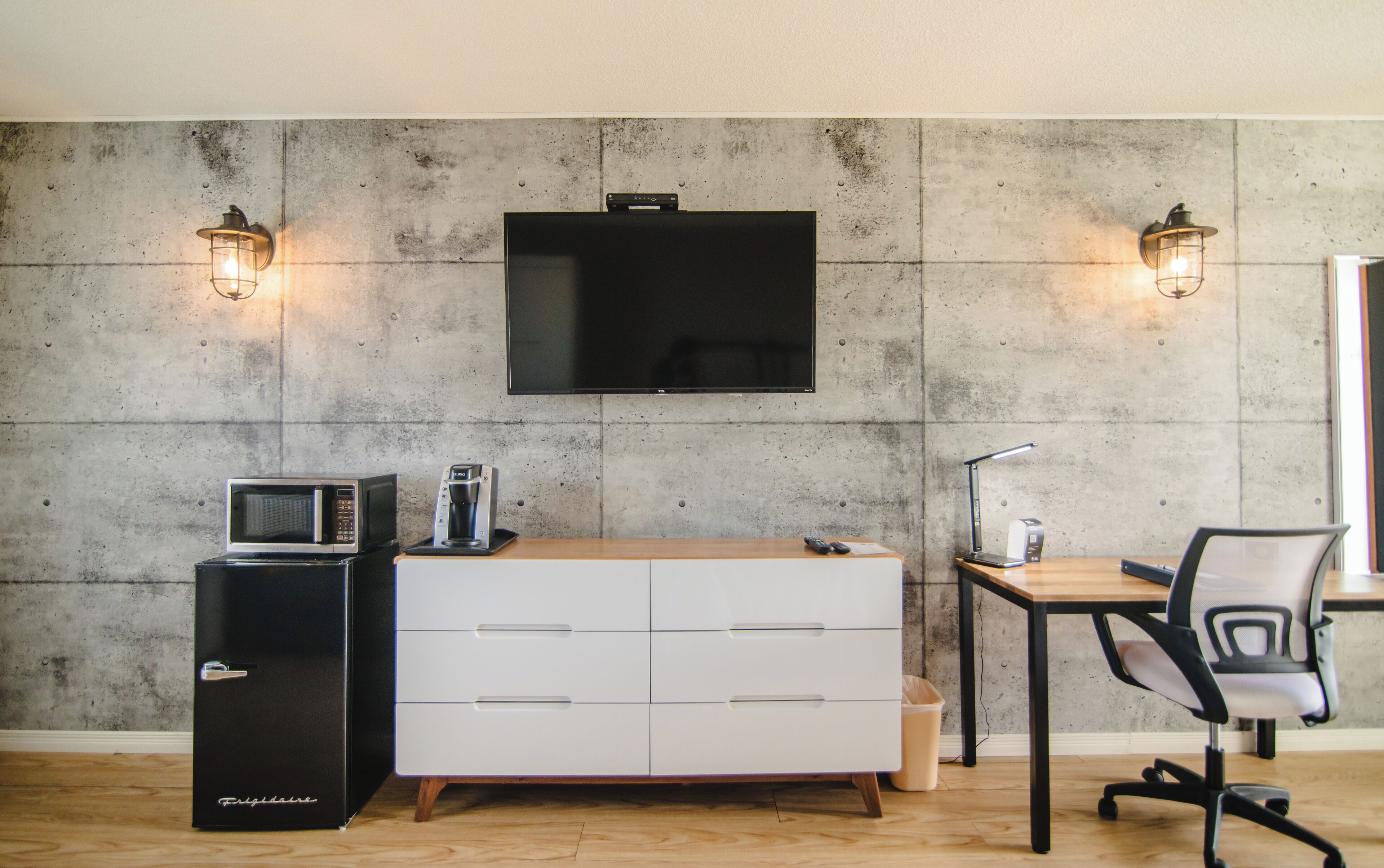 A modern room with a flat-screen TV mounted on a concrete wall, a white dresser with drawers, a small black microwave, a coffee maker, a desk with a lamp, and a black office chair.