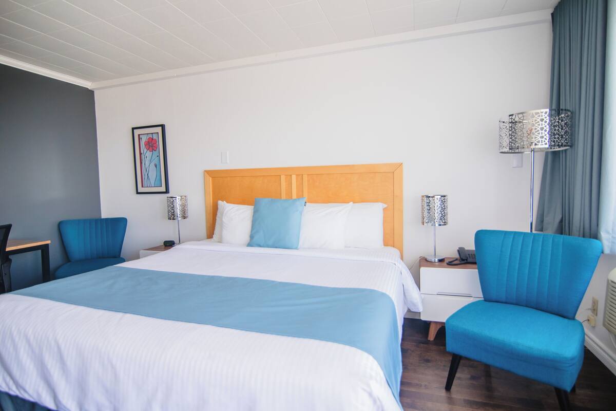 Habitación de hotel con una cama grande, dos sillas azules, una mesita de noche, una lámpara y un cuadro en la pared.