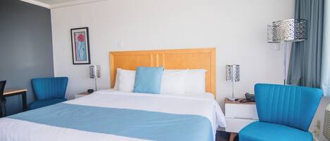 A hotel room with a large bed, two blue chairs, a bedside table, a lamp, and a painting on the wall.