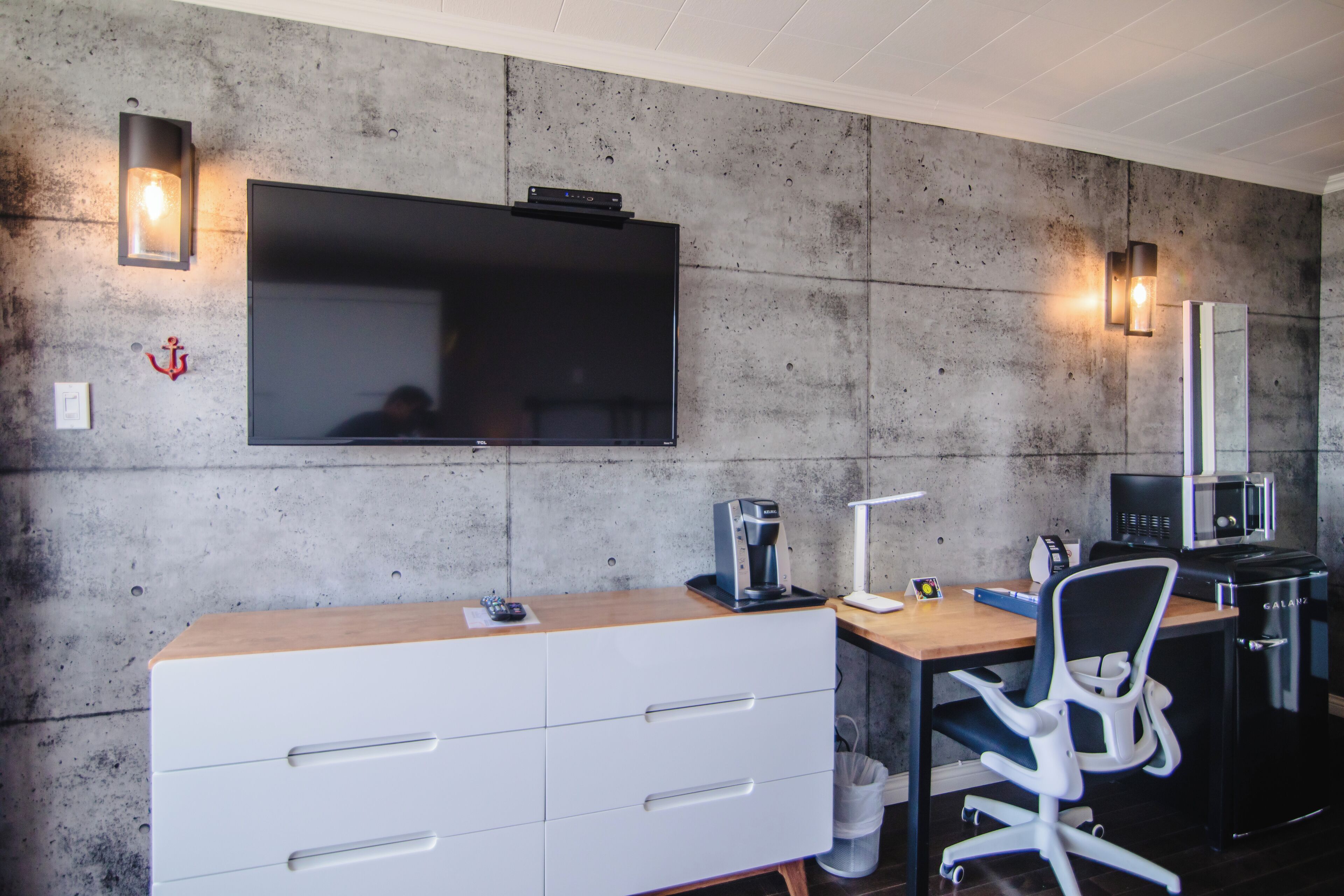 A modern office space with a large flat-screen TV mounted on a concrete wall, a wooden desk with a white drawer unit, a black office chair, a coffee machine, and a black trash bin.