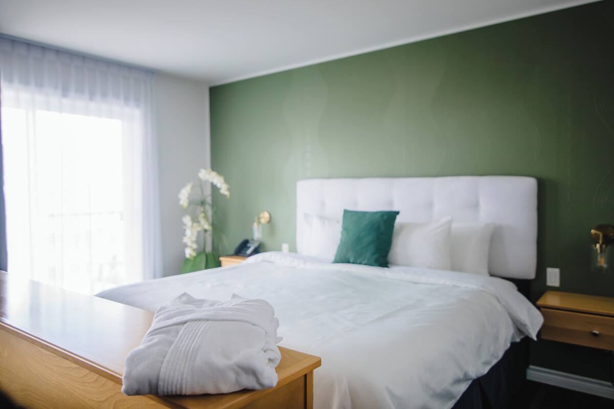 Una habitación de hotel con una cama grande, una mesita de noche de madera, una mesita con una toalla blanca doblada y una pared verde.