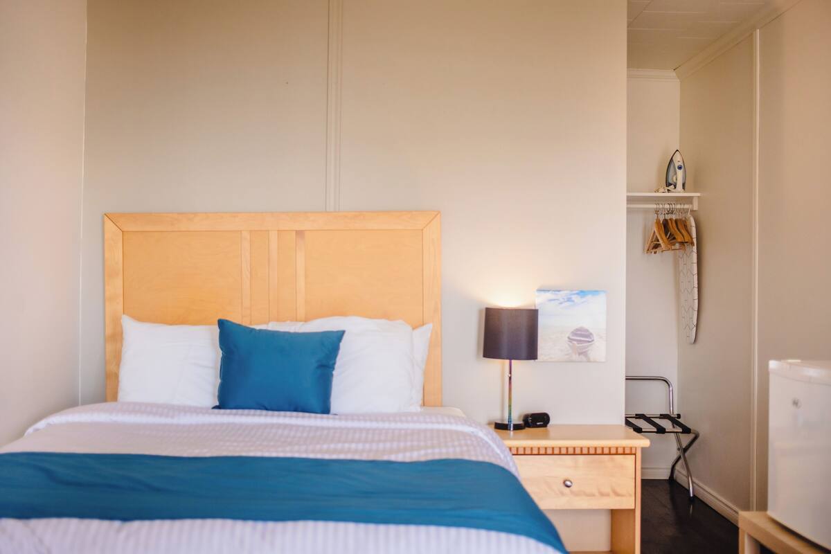 Habitación de hotel con cabecera de madera, cama con ropa de cama blanca y azul, mesita de noche con lámpara, una pequeña mesa y un estante en la pared con un adorno de pez.