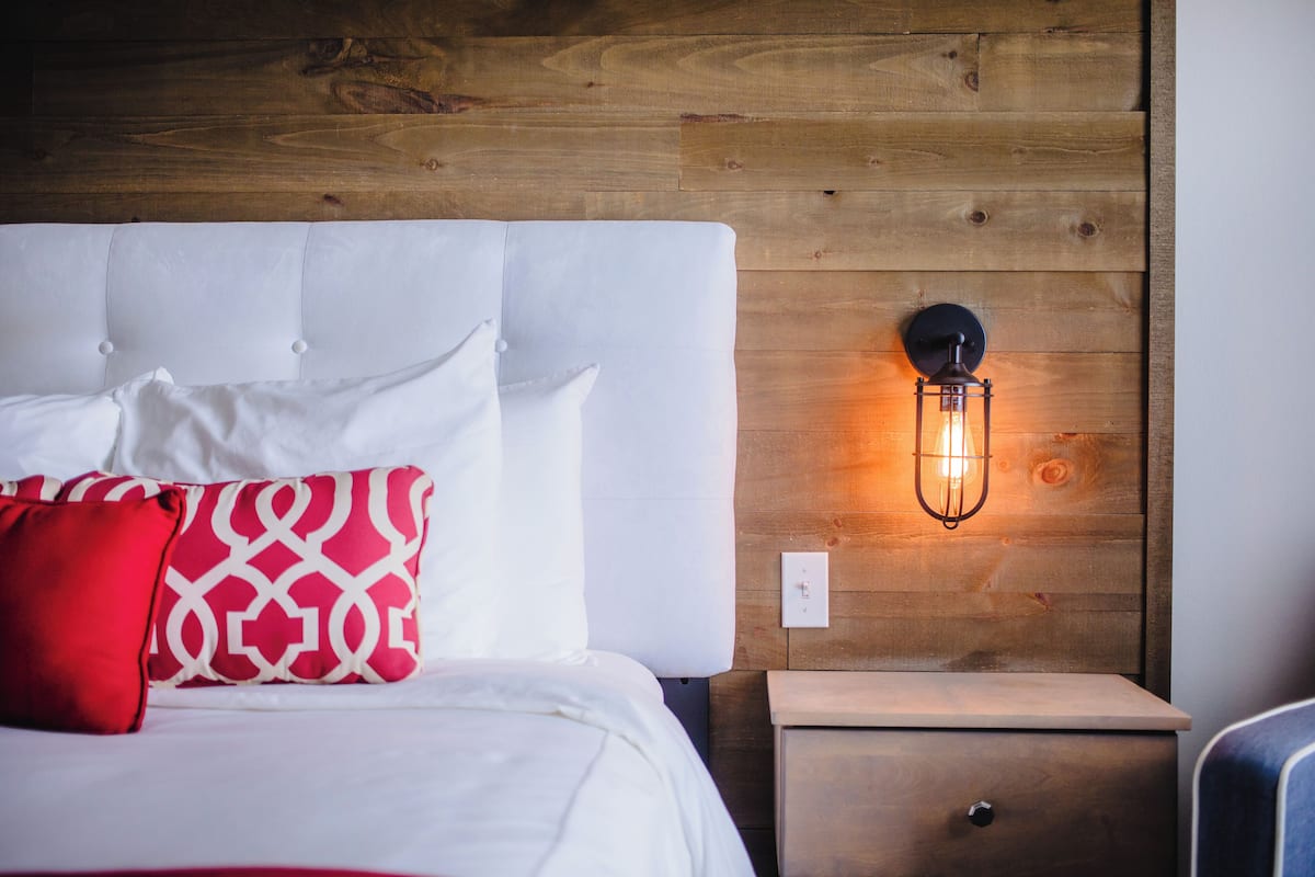 Una cama bien tendida con almohadas blancas y rojas, cabecera de madera y una lámpara fijada a la pared.