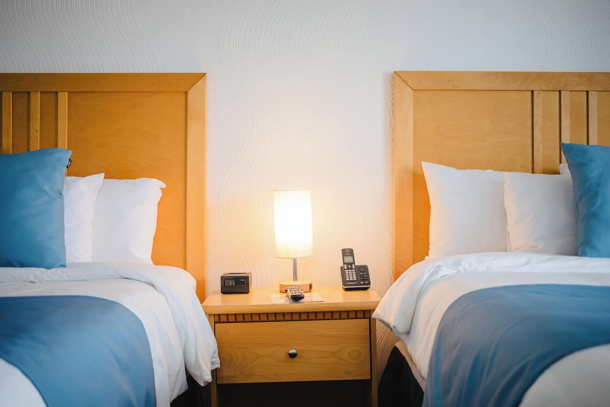 Habitación de hotel con dos camas, cada una con ropa de cama azul y blanca, un cabecero de madera, una mesita de noche con una lámpara, un teléfono y un cajón pequeño.