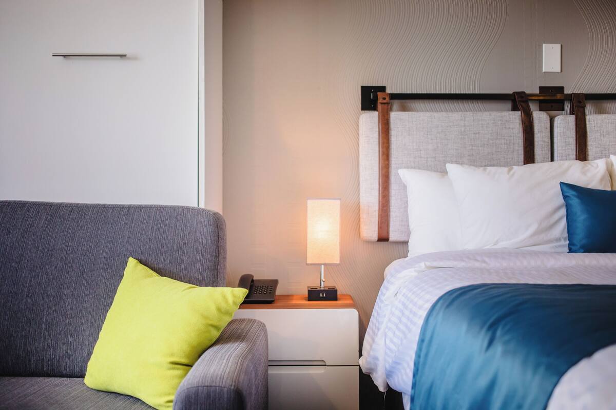 Habitación de hotel con un sofá gris, una mesita de noche con lámpara, teléfono y cama con ropa de cama blanca y azul.