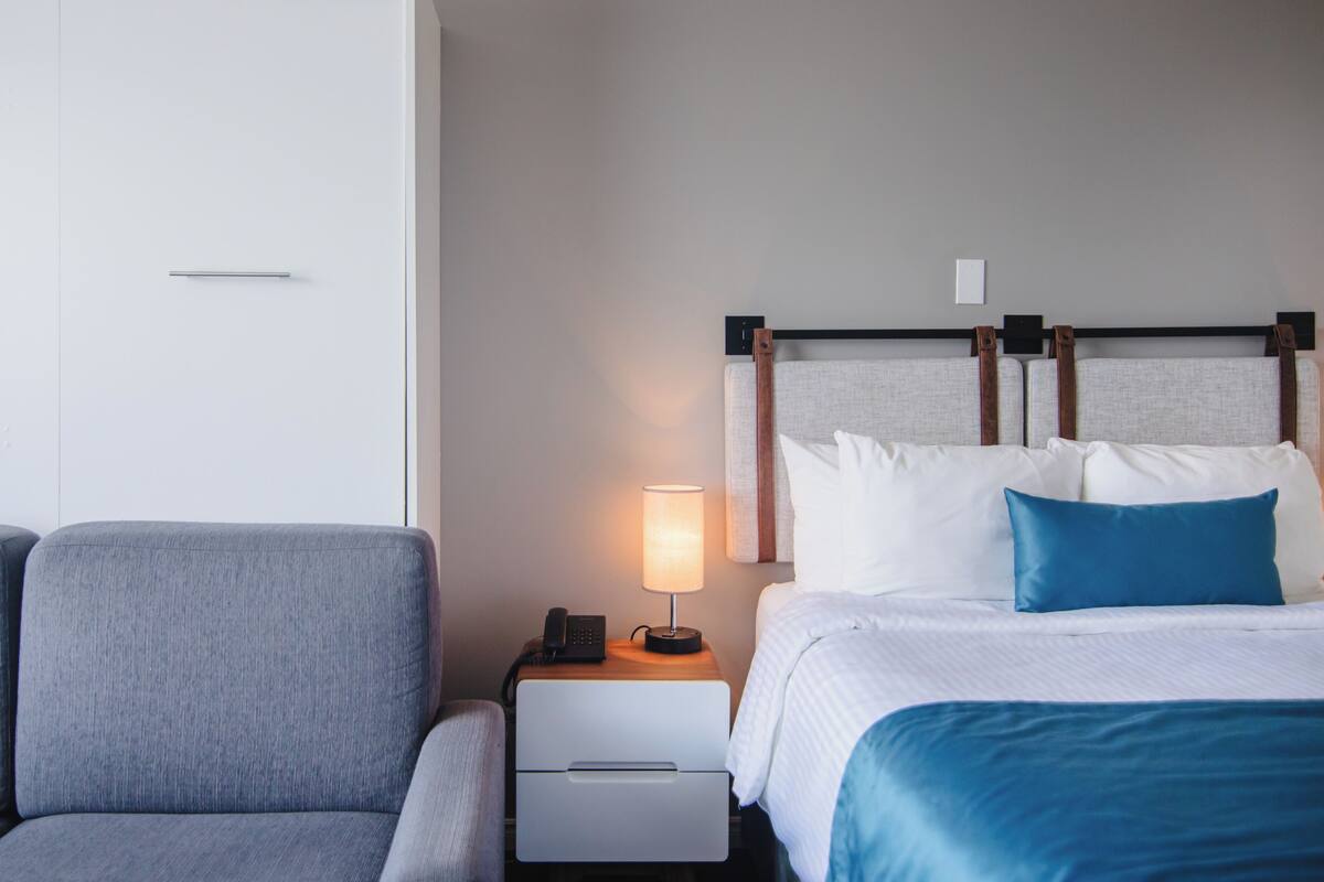Una habitación de hotel con una cama, una mesita de noche con lámpara, un teléfono y un sofá gris.