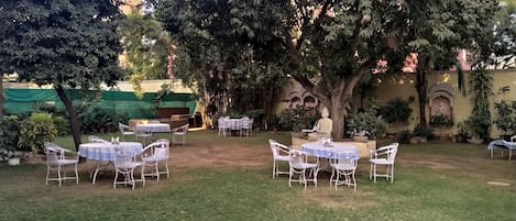 An outdoor seating area with tables and chairs under a large tree, surrounded by potted plants and a decorative statue.