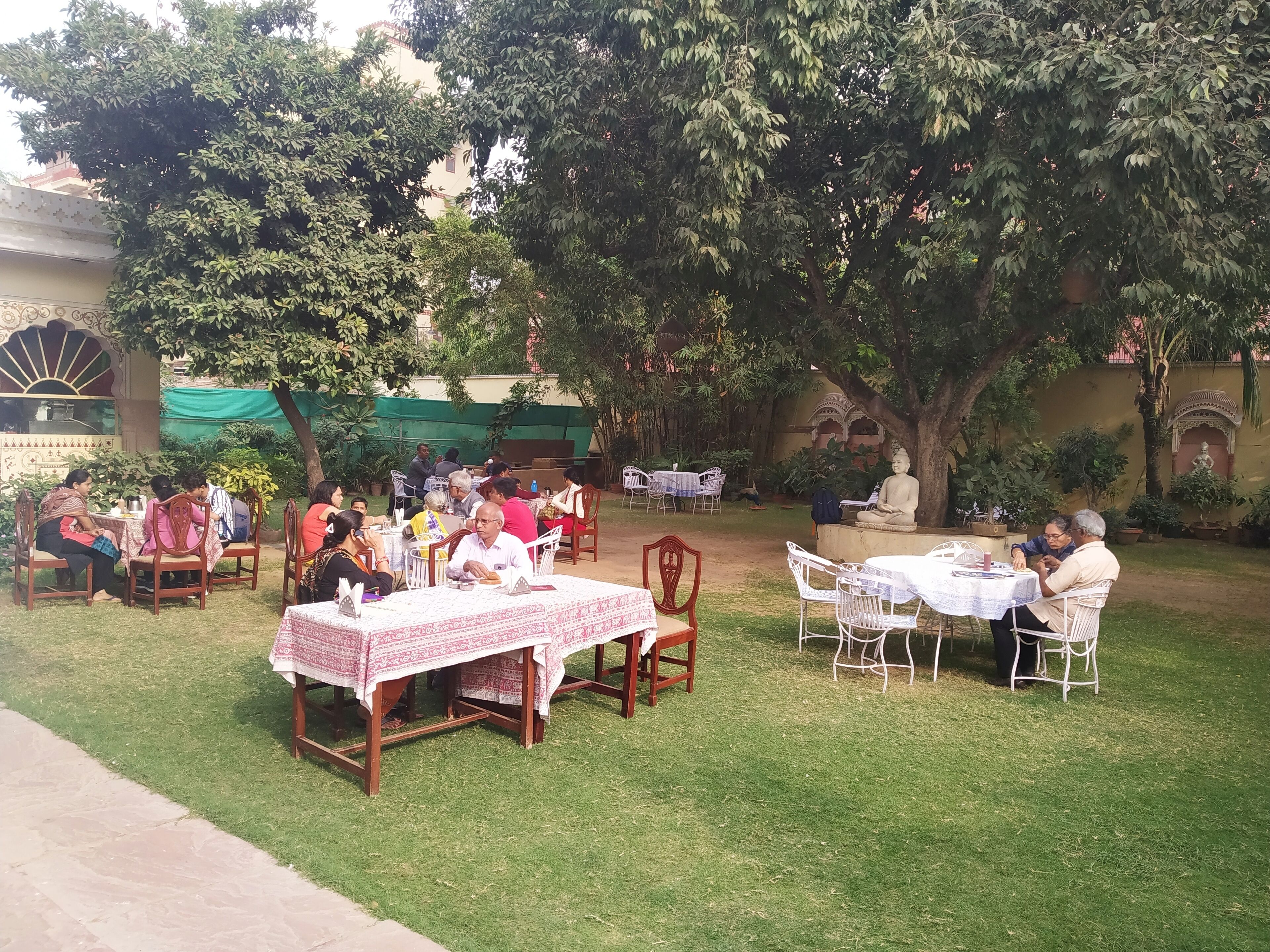 Un área de comedor al aire libre con mesas y sillas, rodeada de follaje y una estatua.