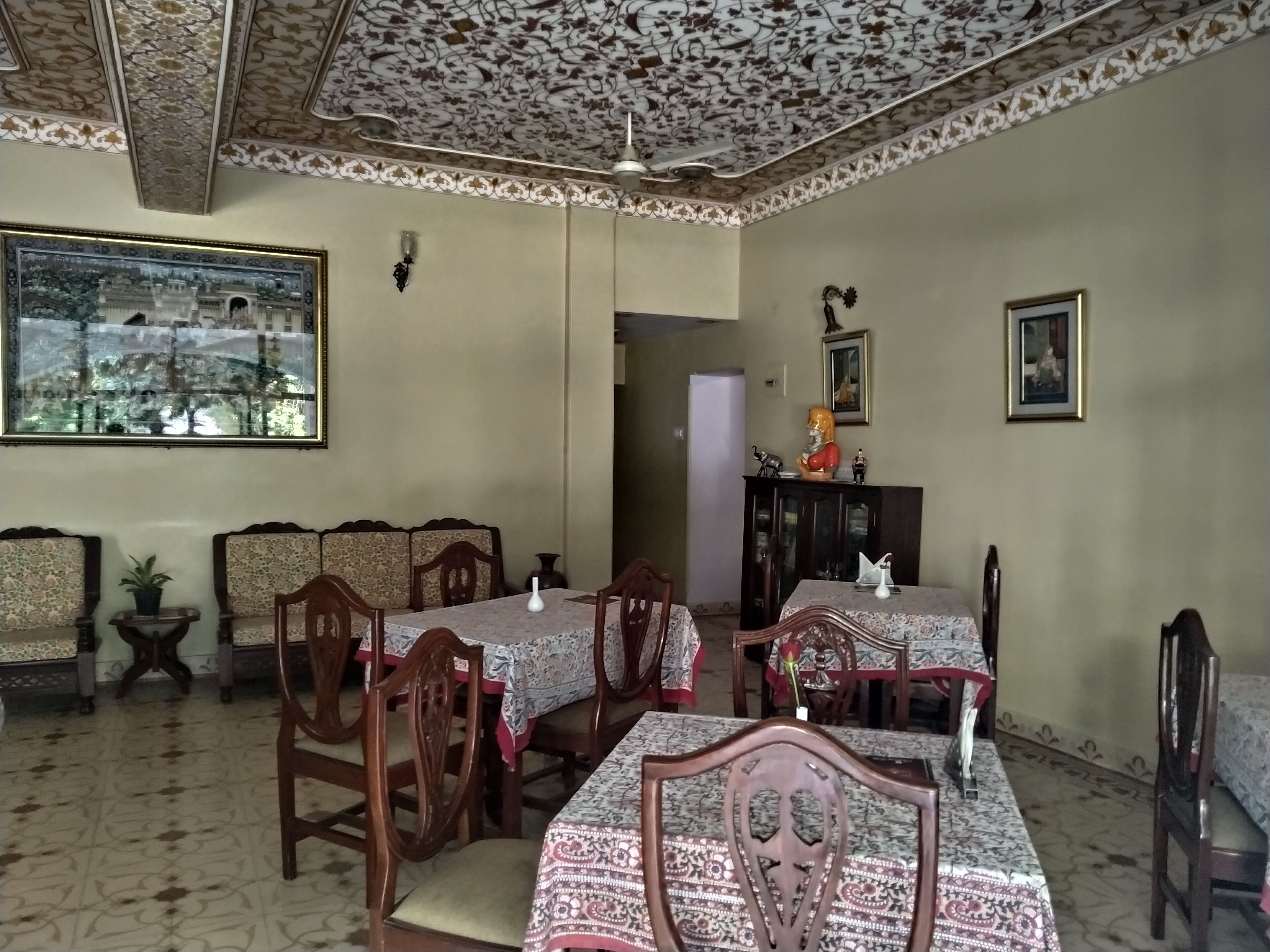 A dining area with wooden chairs and tables, a ceiling with floral patterns, and a large window with a view of buildings.
