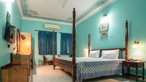 A four-poster bed with a wooden frame and a blue patterned bedspread, a wooden nightstand with a lamp, a wooden bench, a desk with a lamp, and an air conditioning unit on the wall.