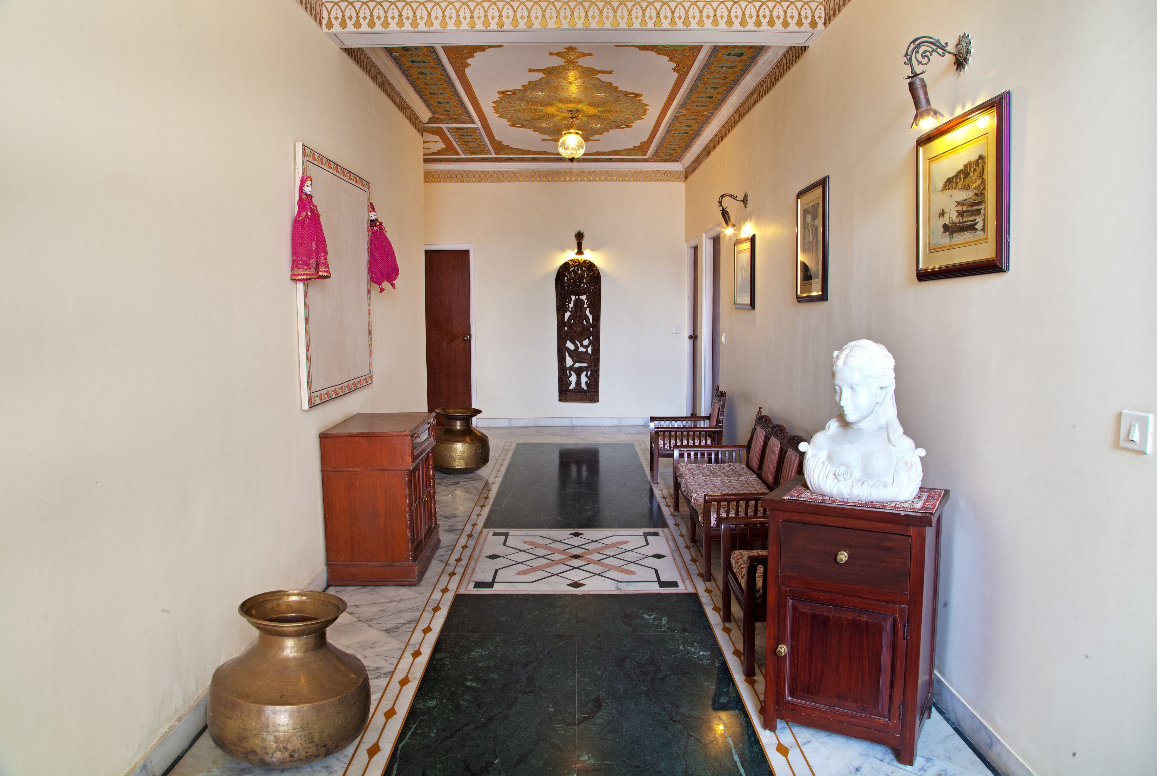 Um corredor com piso de mármore, um banco de madeira, uma escultura de busto e um vaso de latão.