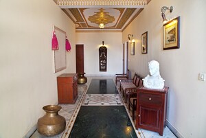 Um corredor com piso de mármore, um banco de madeira, uma escultura de busto e um vaso de latão.