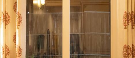 Una habitación con un ventanal grande, cortinas, una silla de madera y una mesa con un florero.