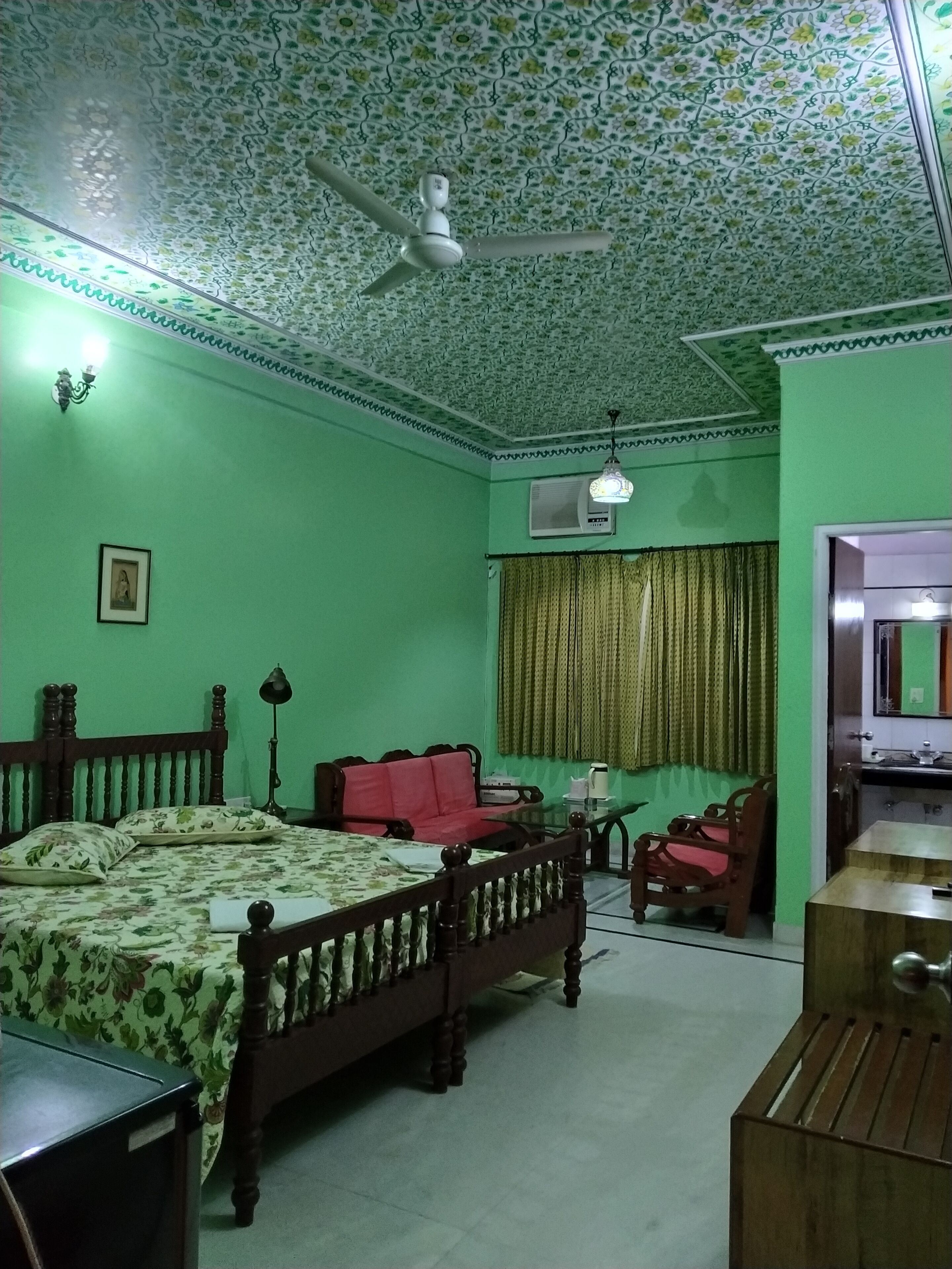 Une chambre à coucher avec un lit à motifs floraux, un canapé rouge, une chaise en bois, un ventilateur de plafond et une petite table avec une lampe.