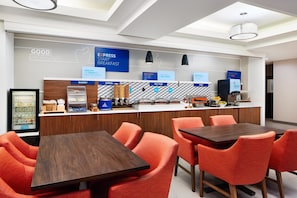 A modern breakfast bar with a variety of food options, including bakery, cereal, and hot food. There are orange chairs and a wooden table in the foreground. - Holiday Inn Express & Suites Wheeling by IHG (Triadelphia)