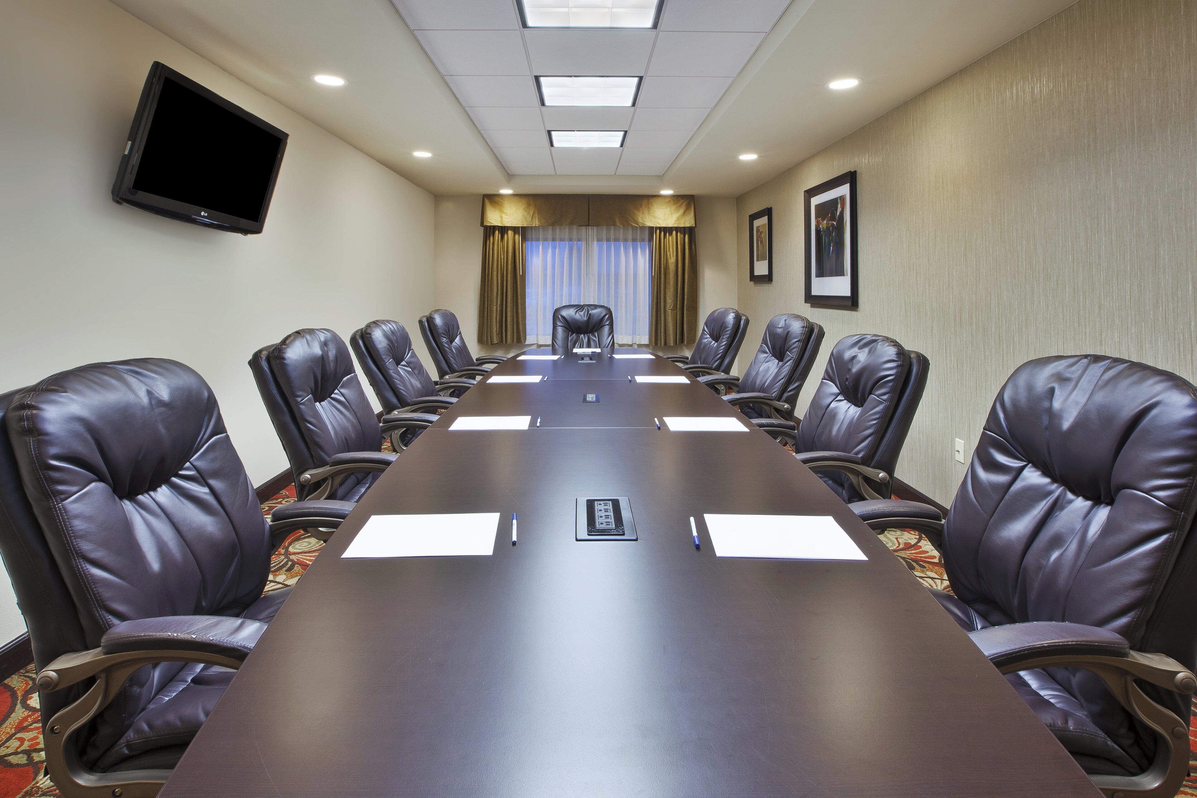 A conference room with a long table, leather chairs, a flat-screen TV, and a window with curtains.