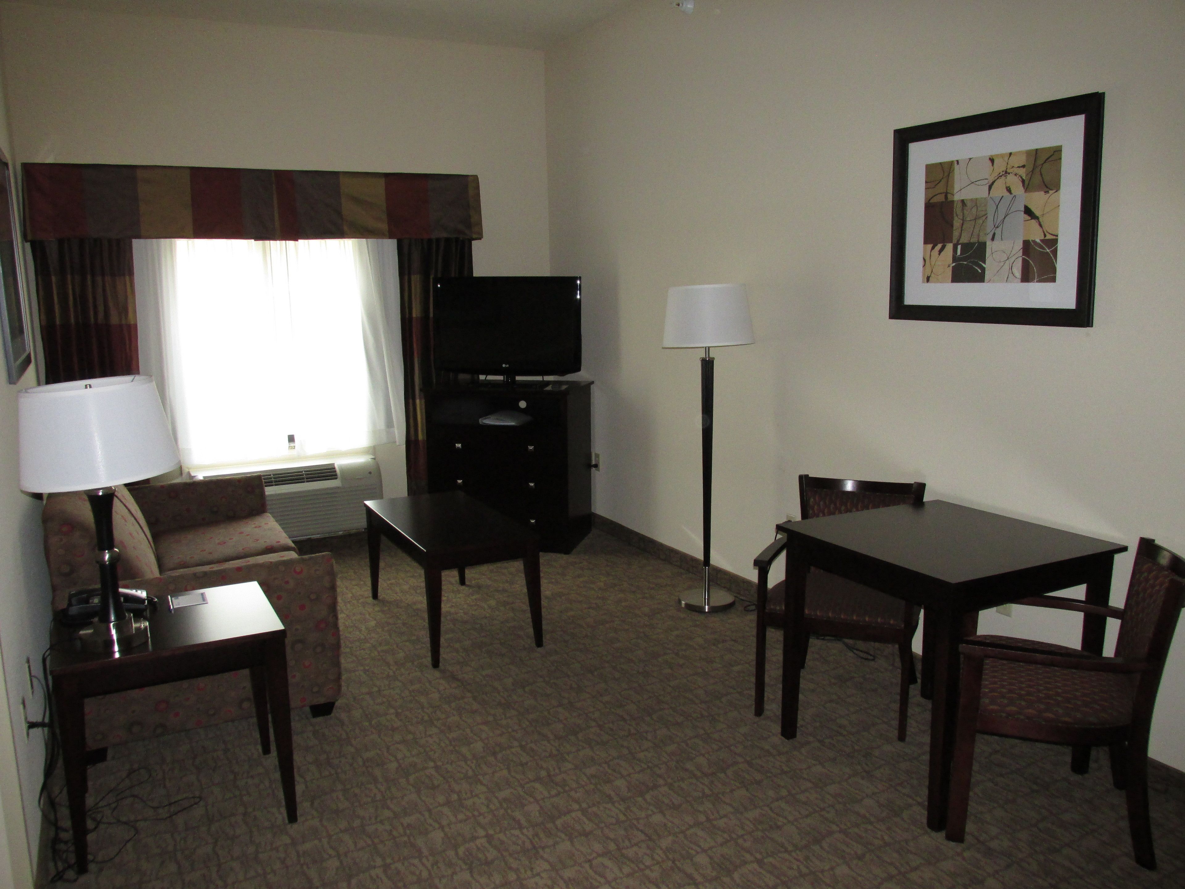 A hotel room with a flat-screen TV, a desk, a chair, a dining table with chairs, a lamp, and a framed picture on the wall.