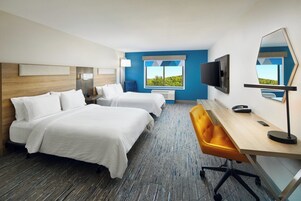 A hotel room with two beds, a desk, a chair, a TV, and a window with a view of greenery.