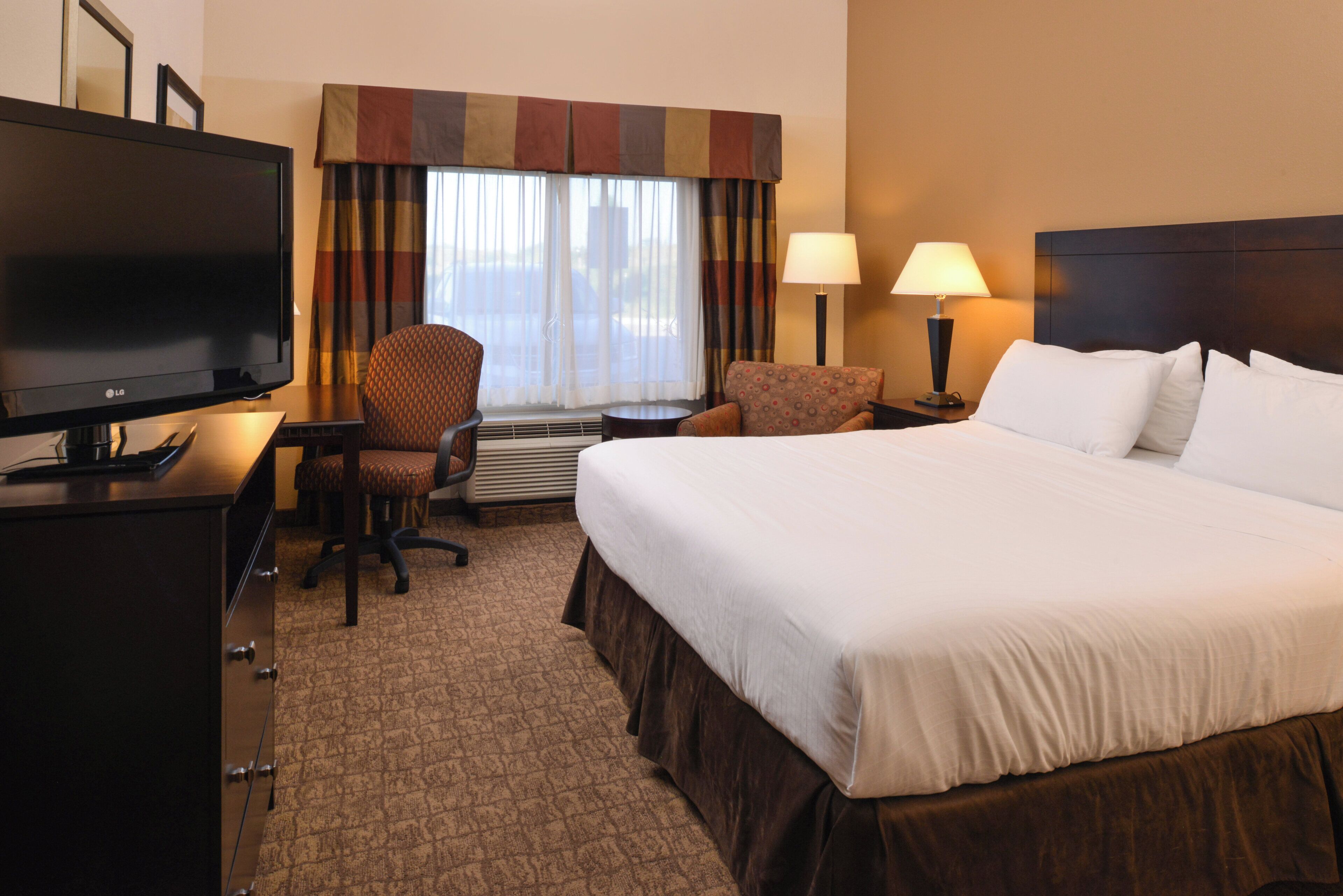 A hotel room with a bed, a desk with a television, two chairs, a window with curtains, and a nightstand with a lamp.
