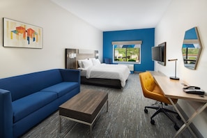 A hotel room with a blue sofa, a bed, a desk with a chair, a TV, and a window with a view of greenery. - Holiday Inn Express & Suites Wheeling by IHG (Triadelphia)