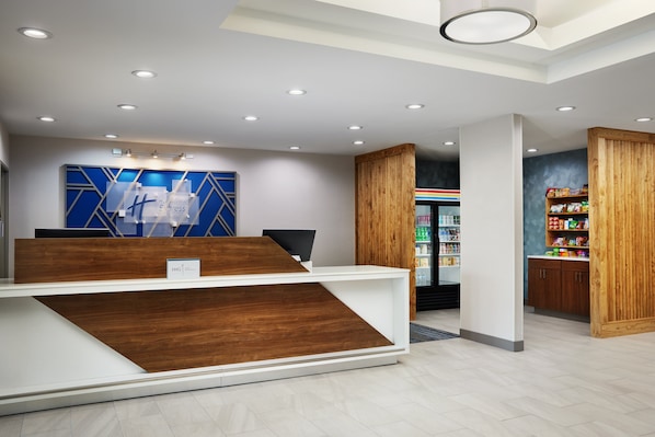 Un área de recepción con un mostrador de madera, un adorno mural azul de diseño geométrico, un refrigerador con refrigerios y un mueble de madera con un cartel colorido.