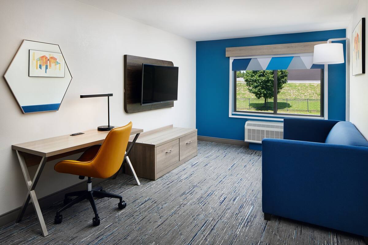 A modern room with a wooden desk, a blue sofa, a mounted TV, and a window with a view of greenery.