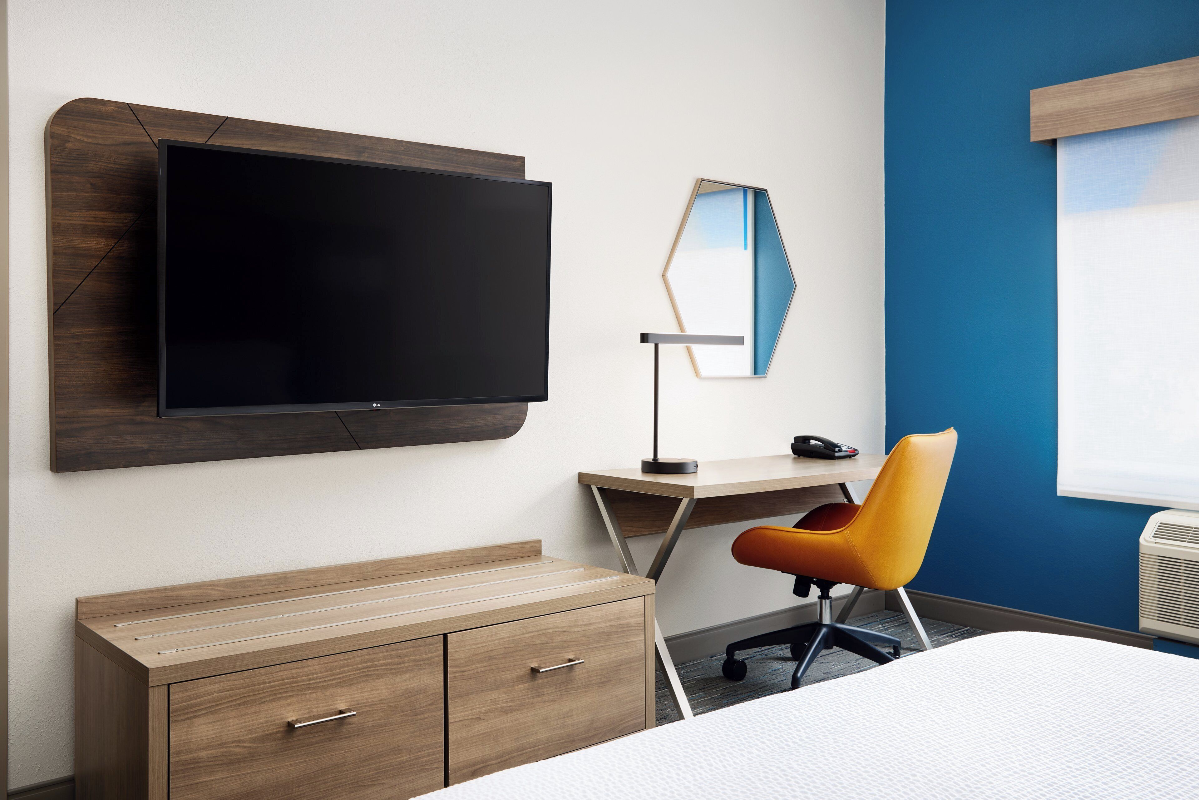 A hotel room with a flat-screen TV mounted on the wall, a desk with a chair, a wardrobe, and a bed.
