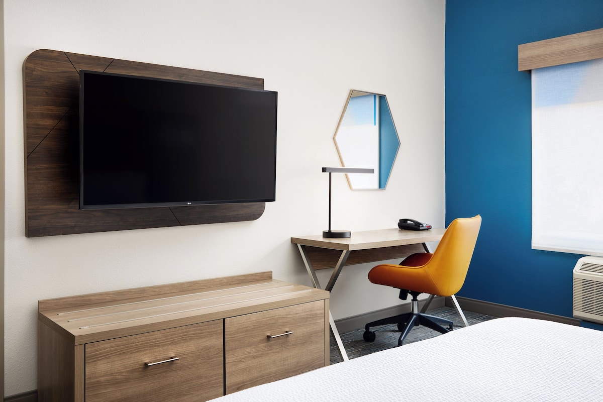 Habitación de hotel con una televisión de pantalla plana en la pared, un escritorio con una silla, un armario y una cama.